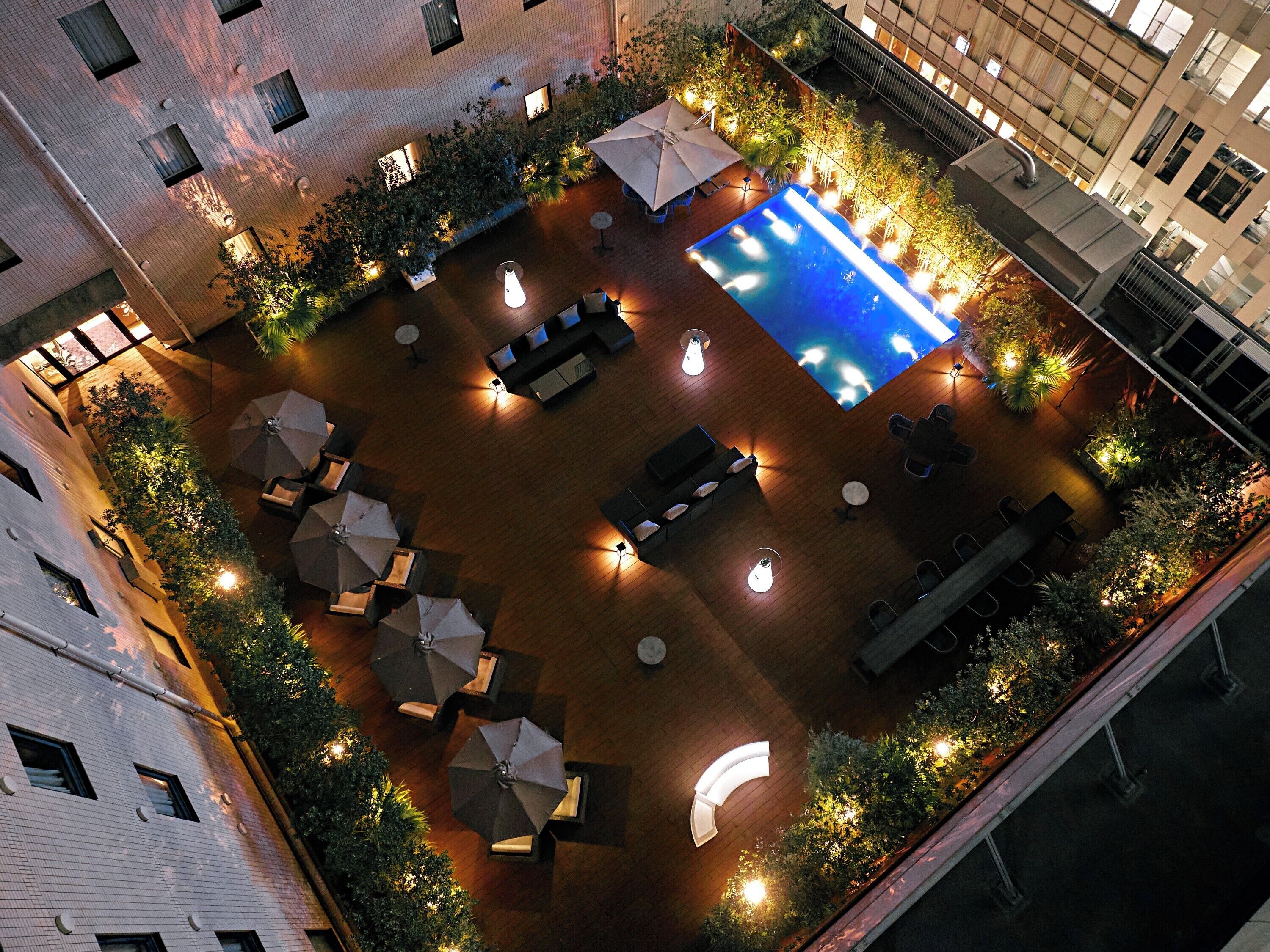 An aerial view of a rooftop pool area with multiple seating arrangements and umbrellas, surrounded by greenery and illuminated by ambient lighting.