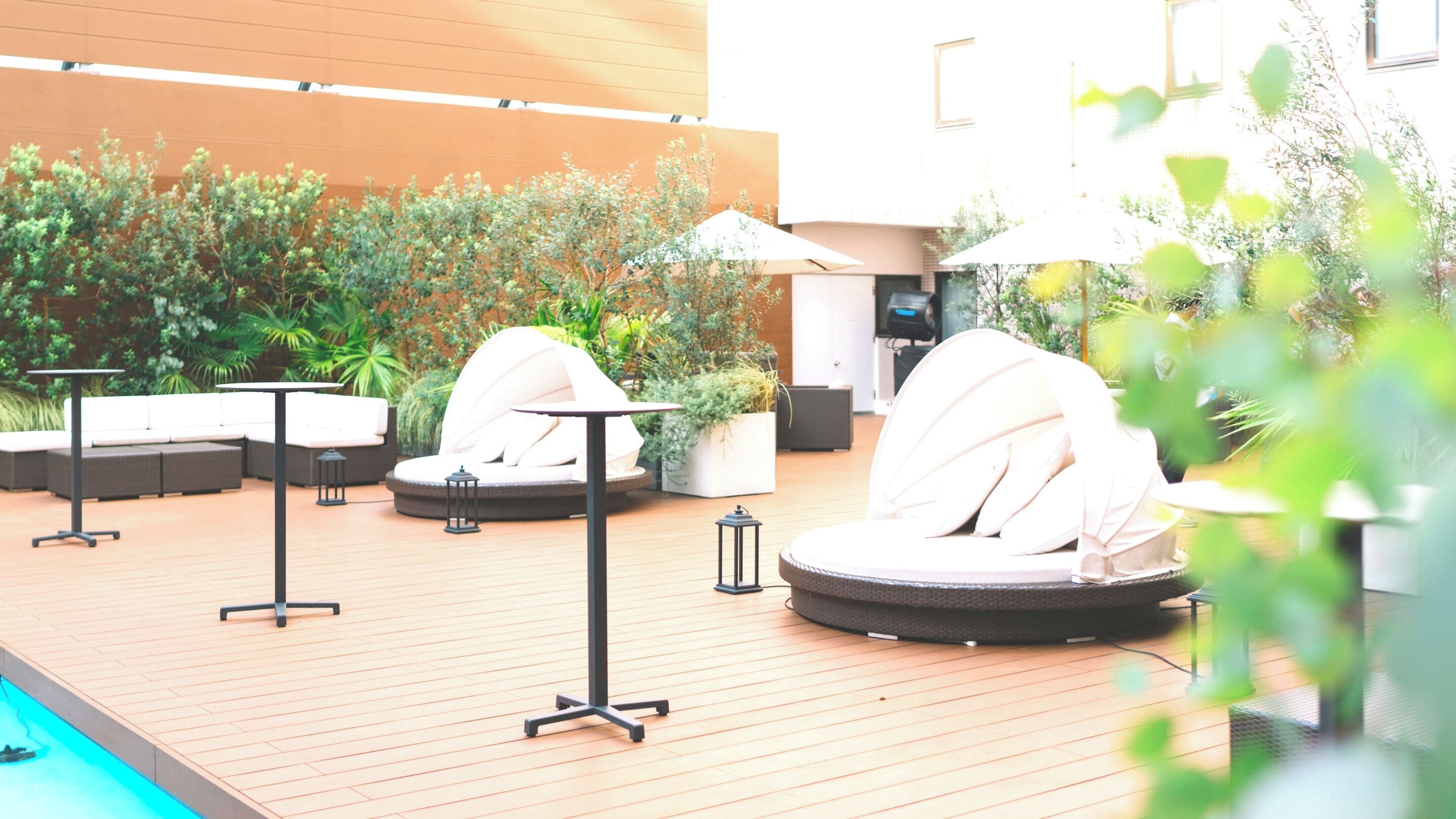 An outdoor seating area with round tables and cushioned chairs, surrounded by greenery and a pool.