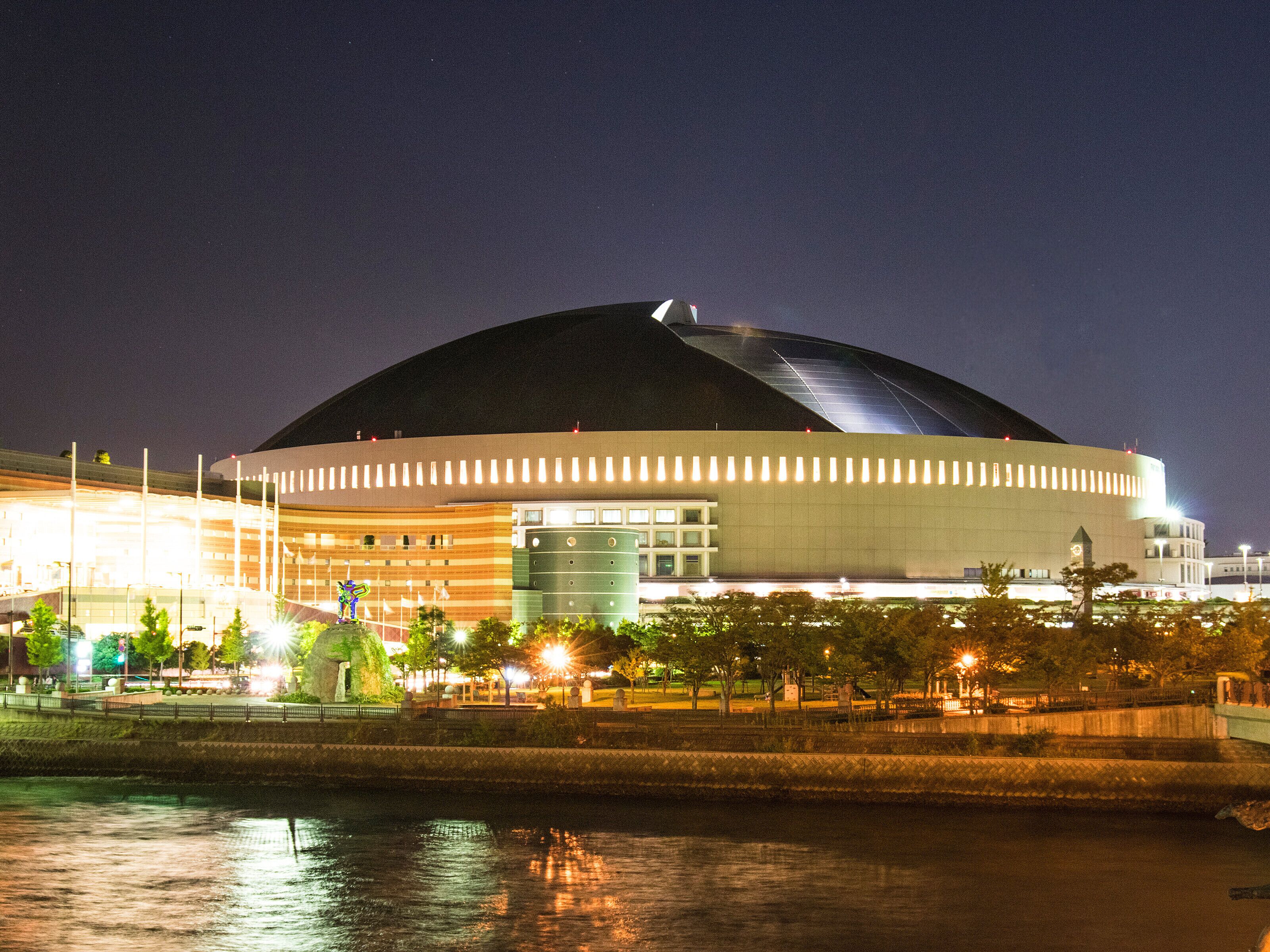 A large, modern stadium with a curved roof, surrounded by a waterfront promenade and illuminated by street lamps.
