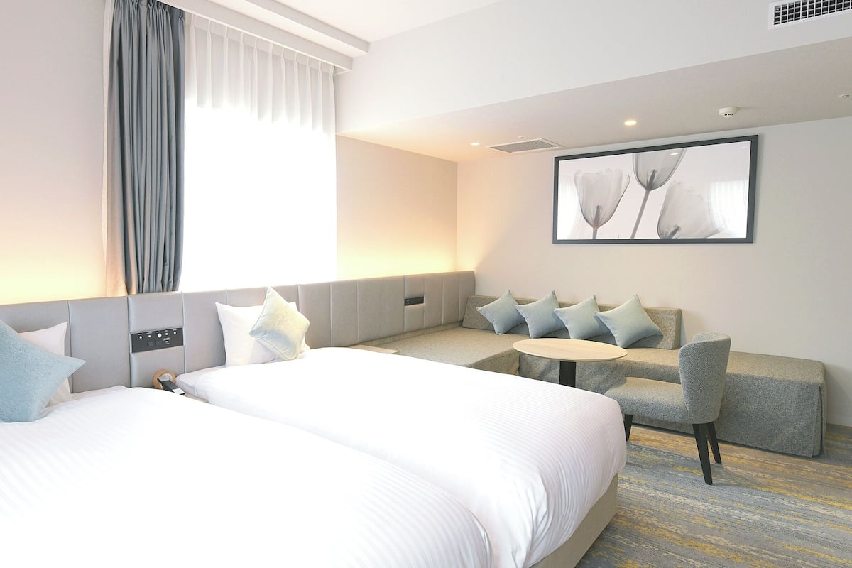 A hotel room with two beds, a sofa, a small table, and a large window with curtains.