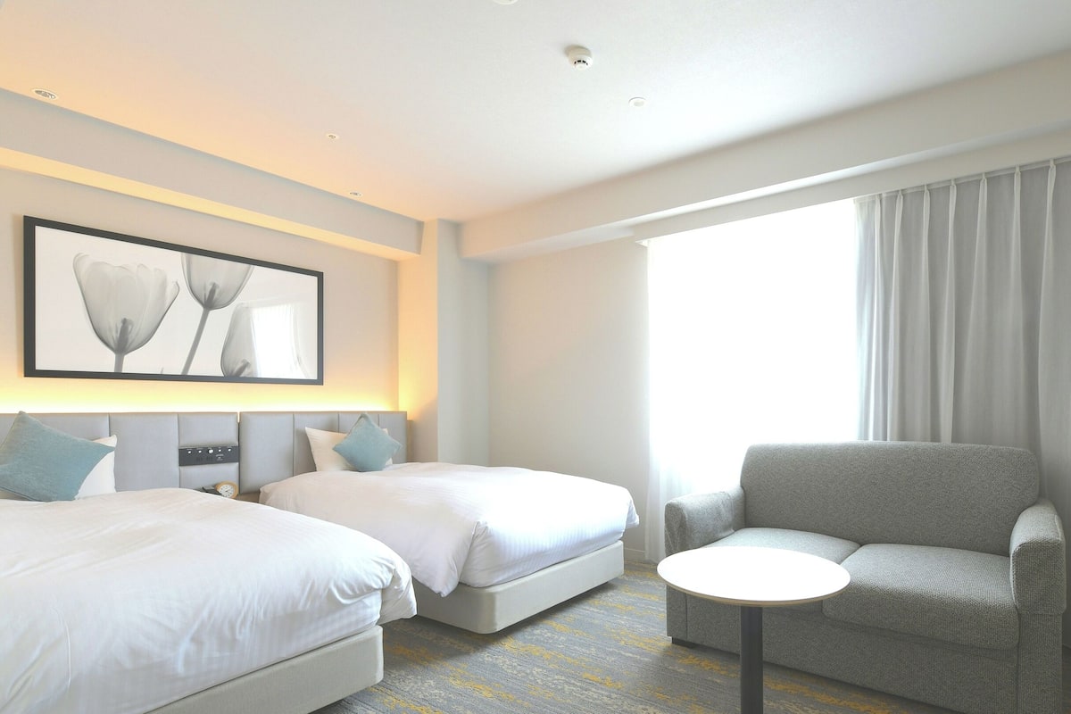 A hotel room with two beds, a gray armchair, a small round table, and a large window with curtains.