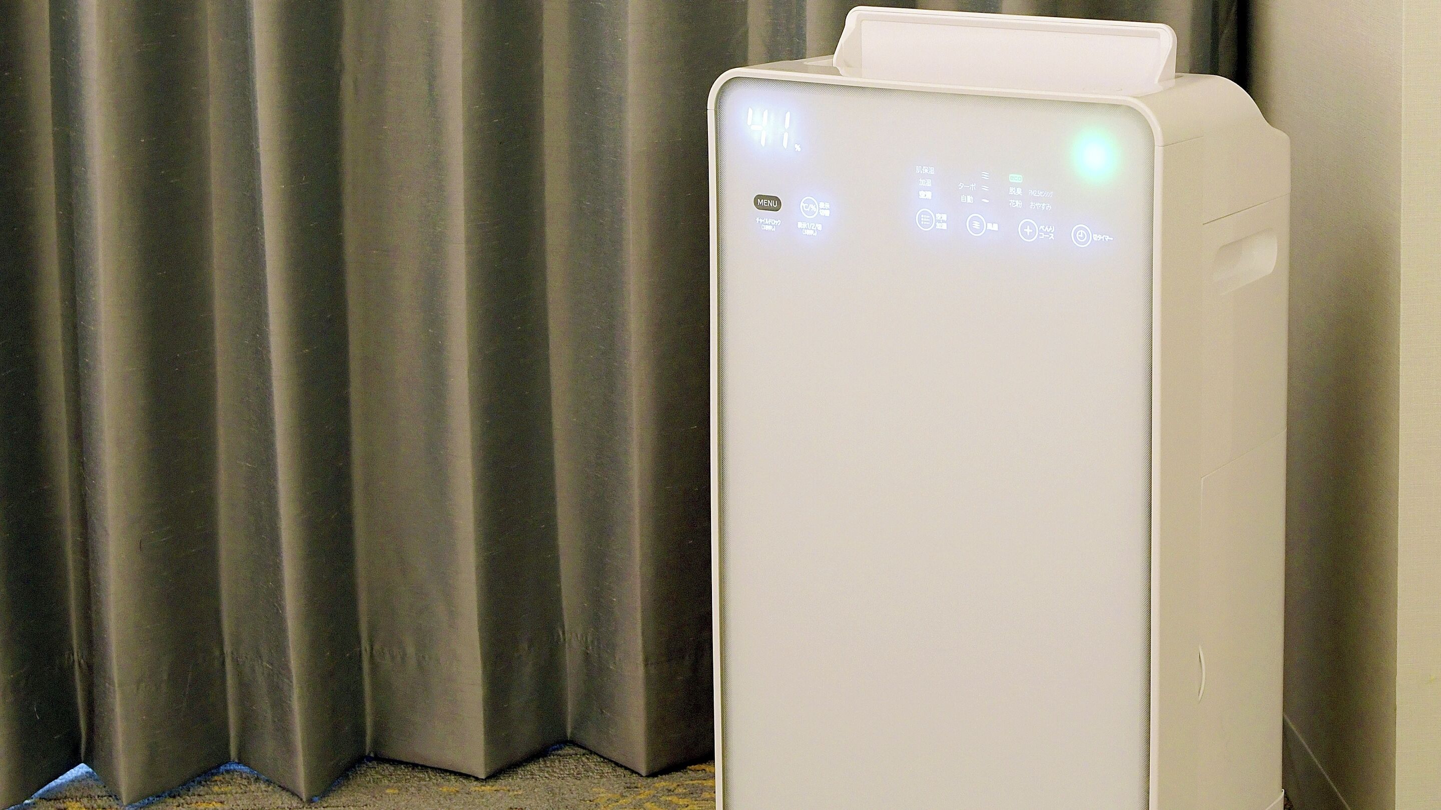 A white air purifier with a digital display, placed near a curtain.