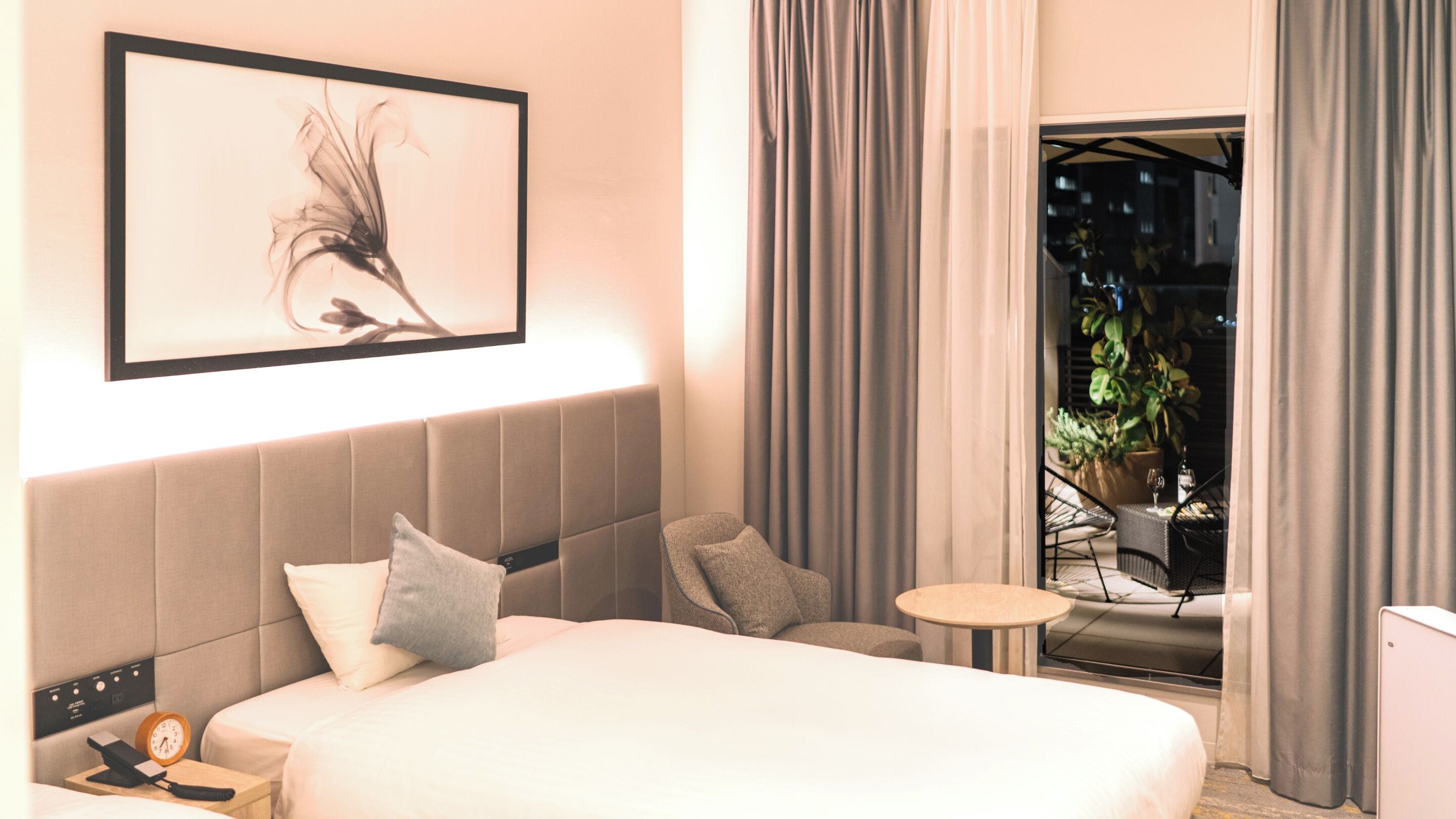 A hotel room with a large bed, a bedside table with a phone and clock, a small round table, and a view of the outside balcony with curtains.