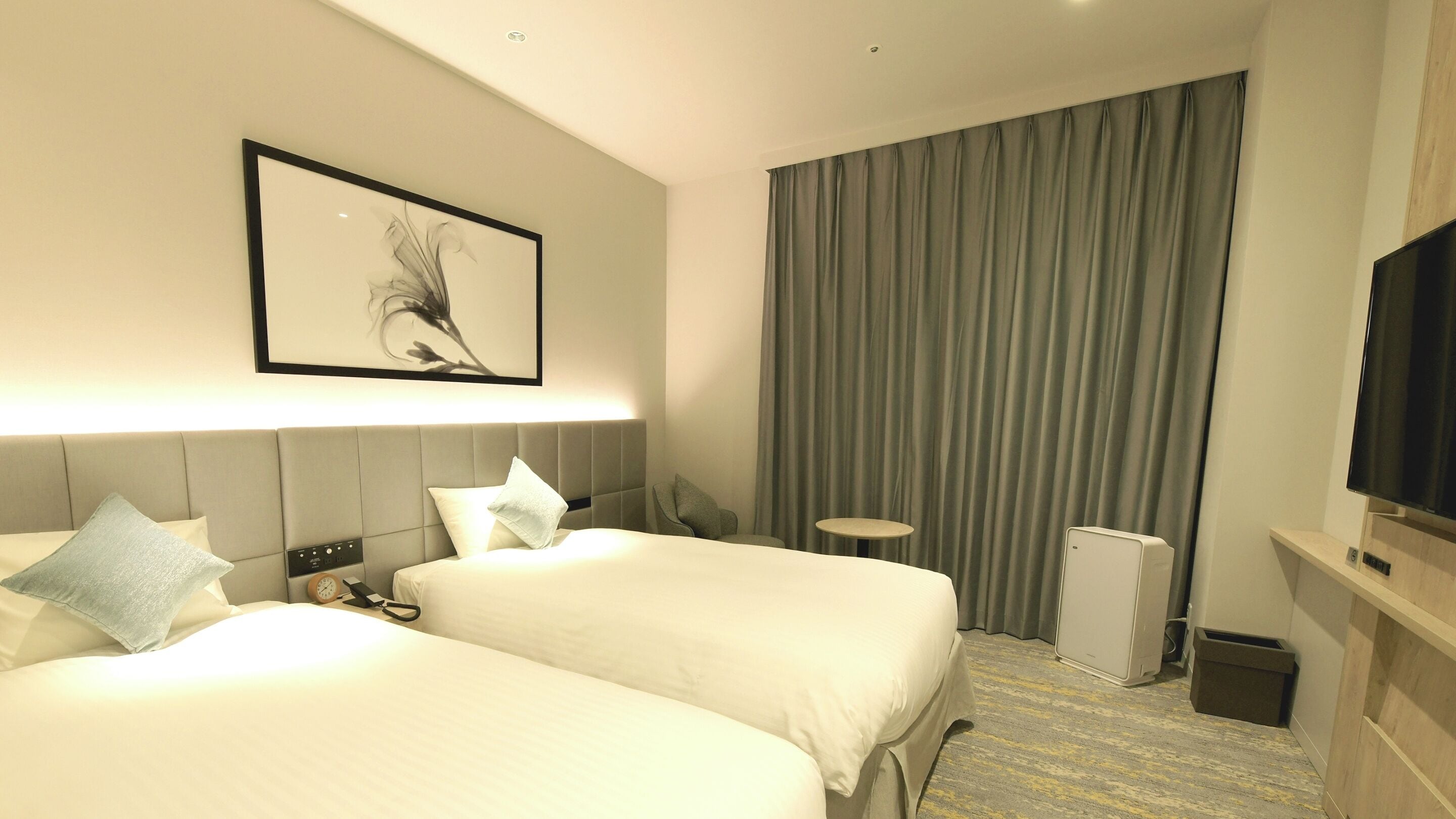 A hotel room with two beds, a flat-screen TV, a small table, a heater, and a curtain.