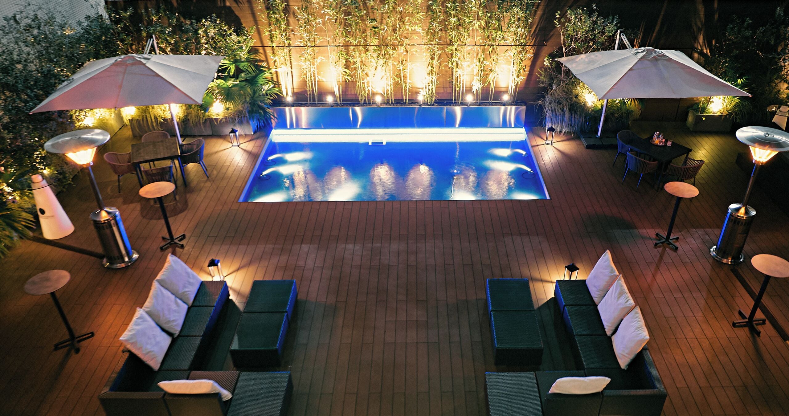 Une piscine entourée de plantes et dotée d’une terrasse en briques, avec des chaises longues et des parasols.