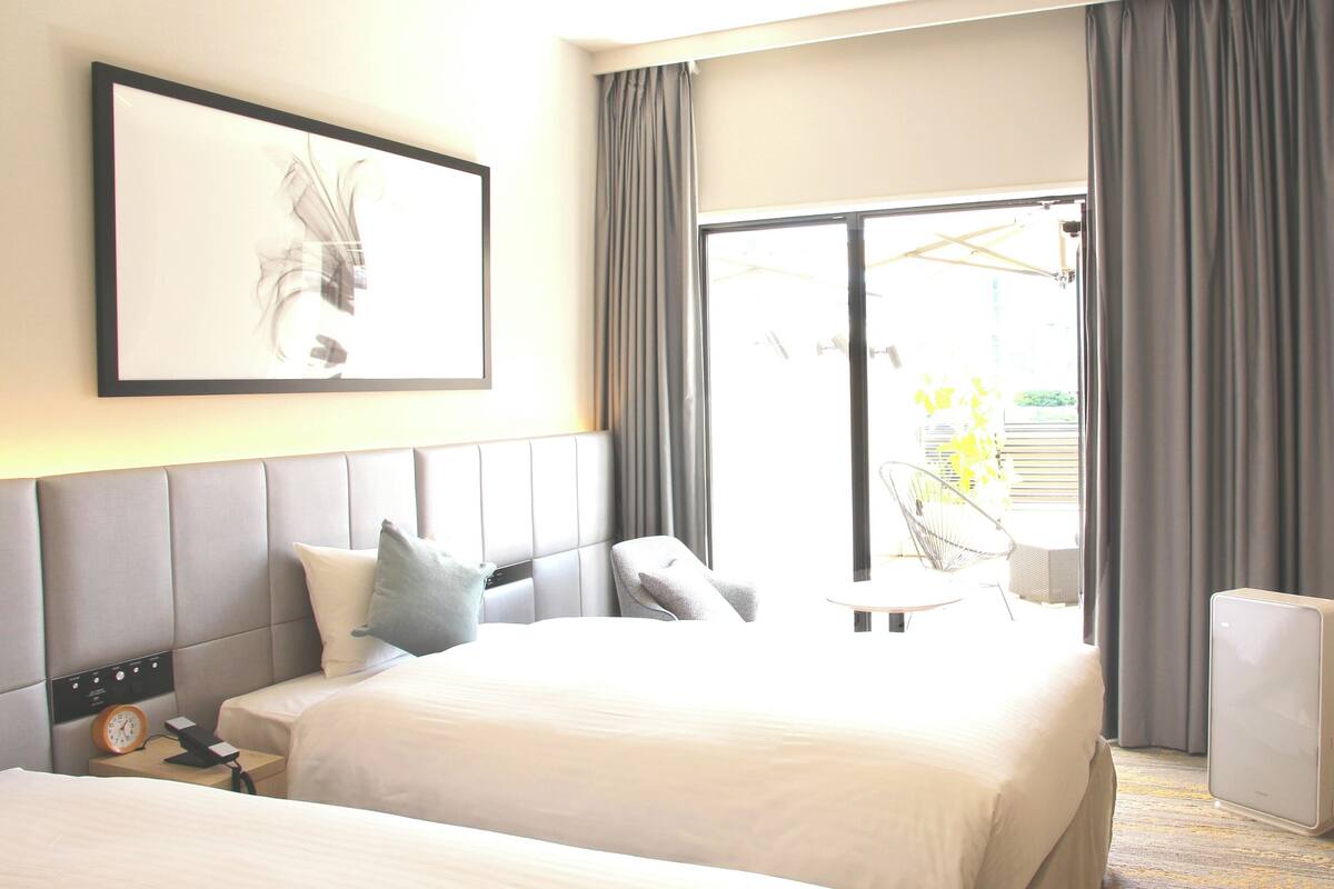 A hotel room with two beds, a large window with curtains, a flat-screen TV, and a framed artwork on the wall.