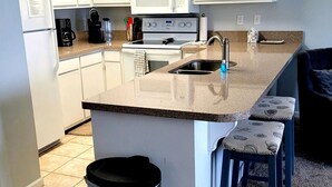Una cocina moderna con armarios blancos, un lavamanos de acero inoxidable, un microondas y una estufa. Hay dos taburetes de bar y un cubo de basura.