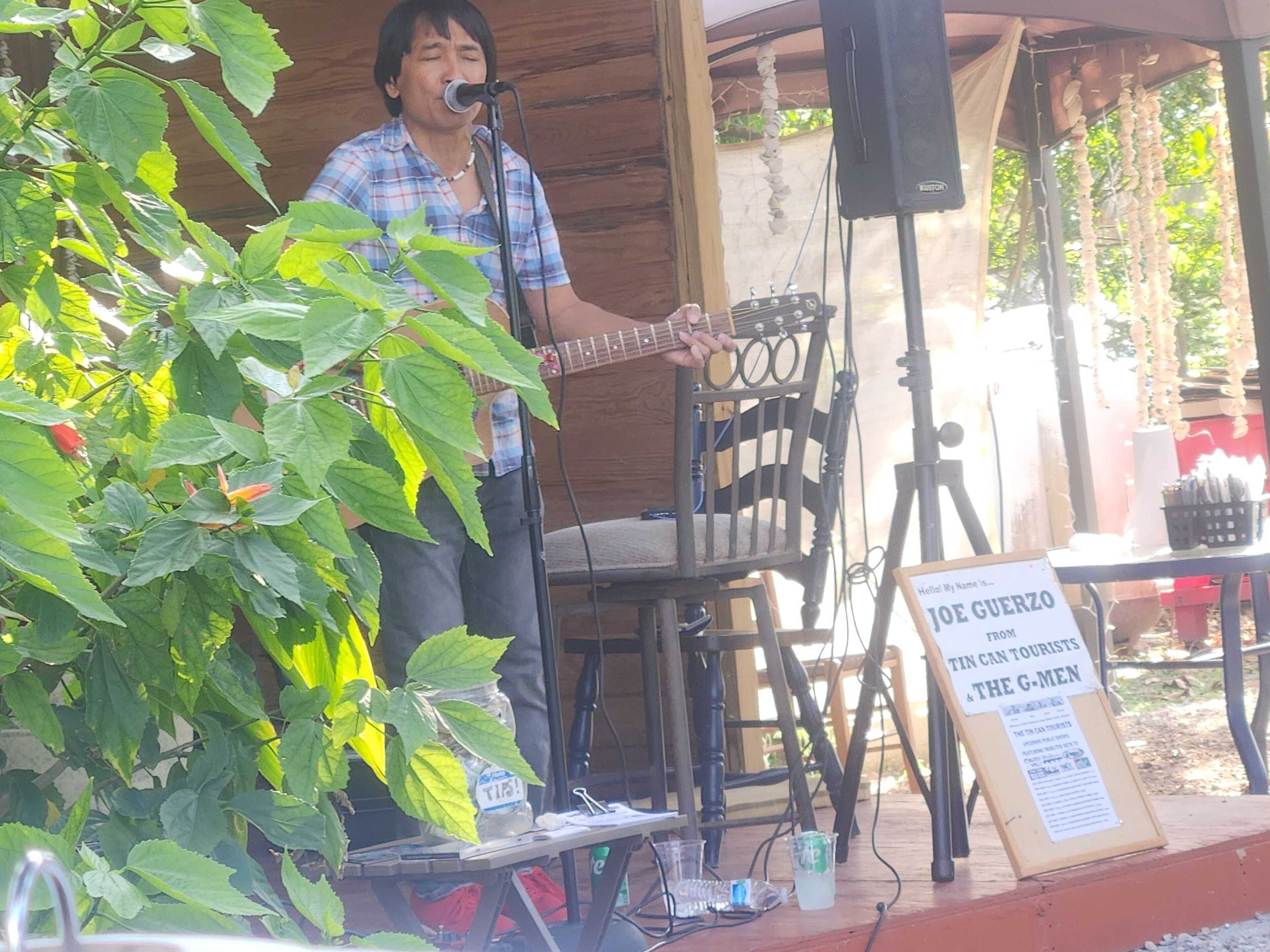 A musician performing on an outdoor stage with a microphone and guitar. There is a signboard with text, and the stage is set up with chairs and tables.