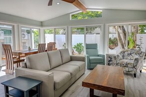 A spacious living area with a sofa, a coffee table, and a dining table. There are large windows providing natural light and views of the outdoors.