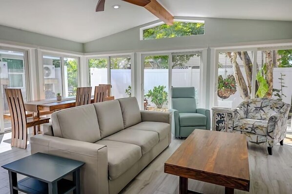 A spacious living area with a sofa, a coffee table, and a dining table. There are large windows providing natural light and views of the outdoors.