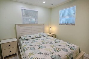 A bedroom with a bed, two bedside tables, a nightstand, and two windows with blinds.