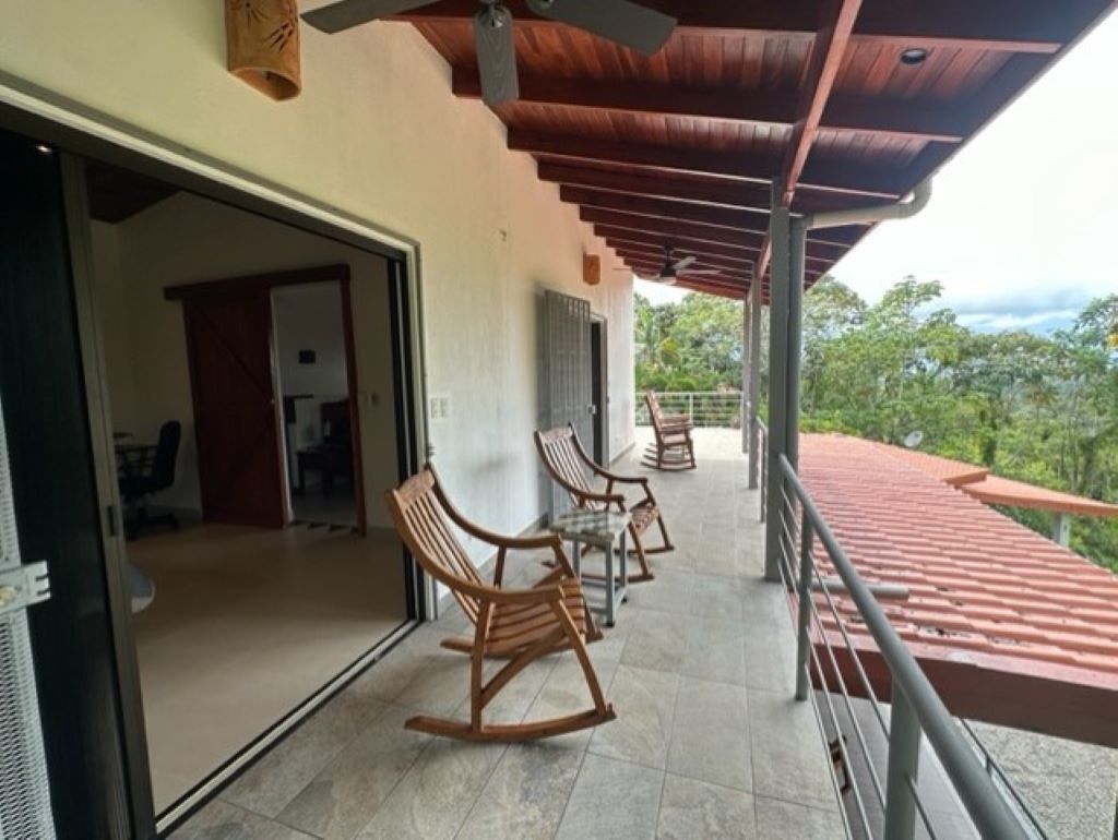 A covered outdoor area with wooden rocking chairs and tables, a door leading to an interior space, and a view of greenery.