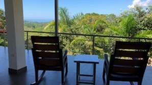 A balcony with two wooden chairs and a small table, overlooking a lush landscape with greenery and a view of the ocean.
