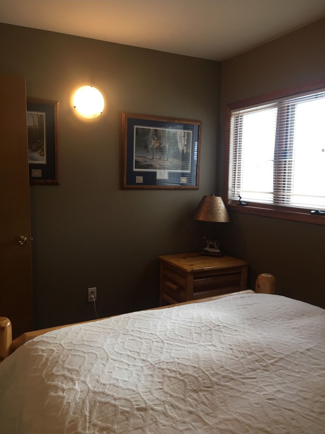 A bedroom with a bed, a nightstand, a lamp, a framed picture, and a window with blinds.