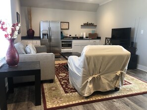 Een woonkamer met een bank, fauteuil, salontafel, vloerkleed, een koelkast, oven, televisie en een vaas met bloemen.