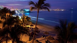 Una playa con palmeras, sombrillas y vista a un paisaje urbano al anochecer.