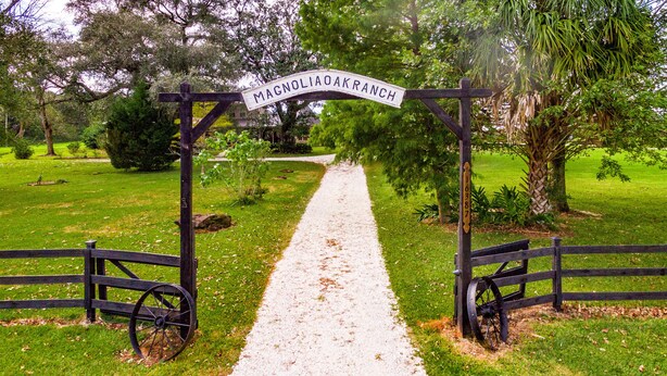 Ein Kiesweg führt zu einem hölzernen Tor mit einem Schild, auf dem "Magnolia Oak Ranch" steht.