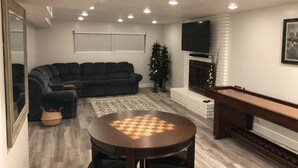 A living room with a dark-colored sofa, a round wooden table with a chessboard, a small Christmas tree, and a fireplace.