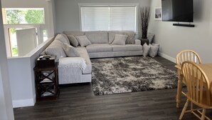 A living room with a grey sectional sofa, a black and white rug, a wooden dining table with chairs, and a TV mounted on the wall.