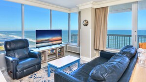 A living room with a large TV, leather sofas, a coffee table, and a view of the ocean.