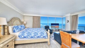 A hotel room with a bed, a desk, a chair, a TV, and a view of the ocean.