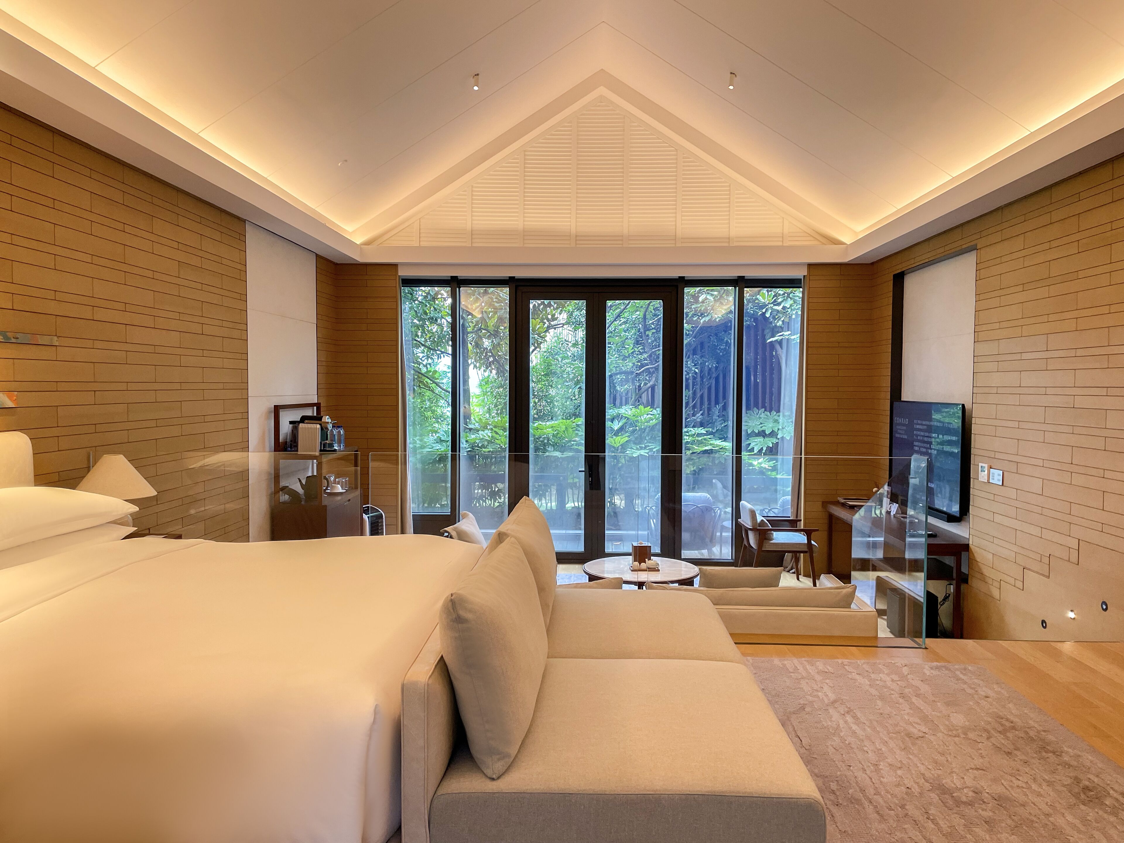 A modern hotel room with a large bed, a sofa, a TV, and a view of a lush garden through large windows.