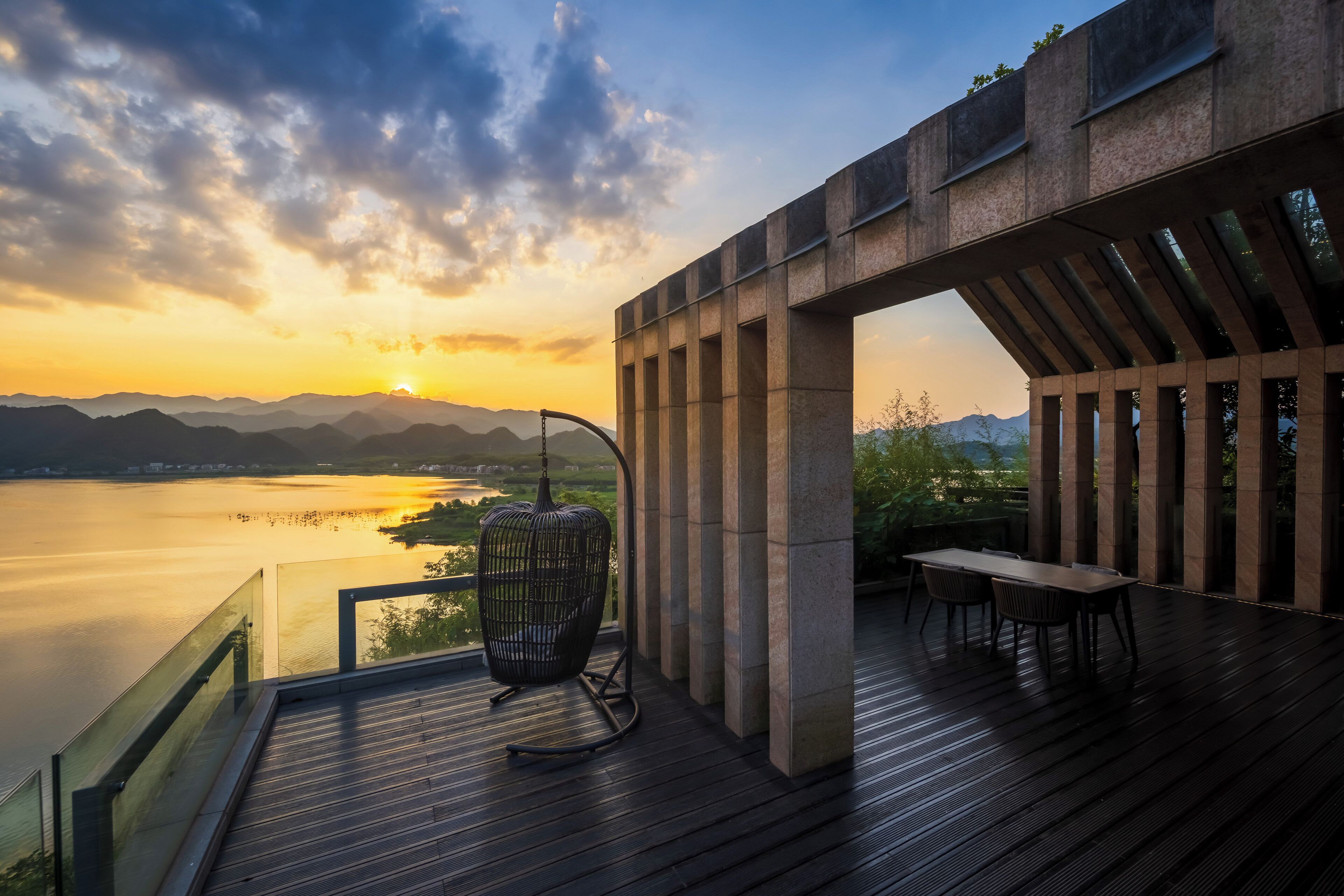 A wooden deck with a glass railing overlooking a body of water and mountains at sunset.