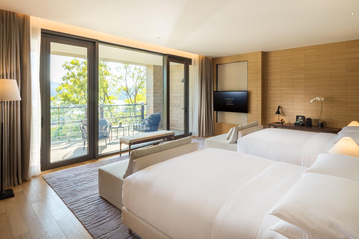 A hotel room with two beds, a TV, a balcony with outdoor furniture, and a wooden wall.