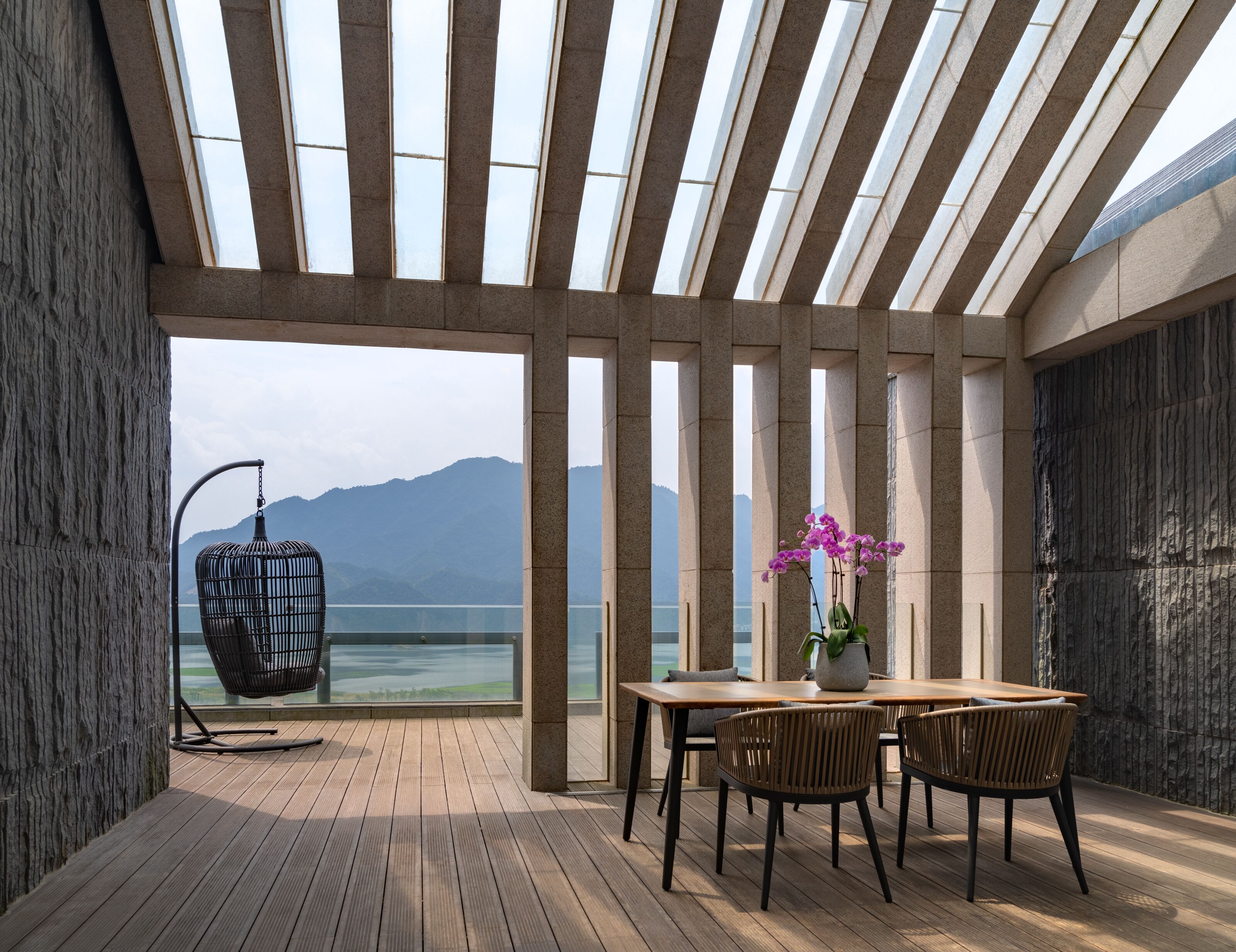 A modern outdoor dining area with a wooden table, chairs, and a hanging chair, overlooking a body of water and mountains.