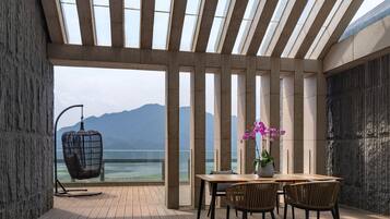 A modern outdoor dining area with a wooden table, chairs, and a hanging chair, overlooking a body of water and mountains.