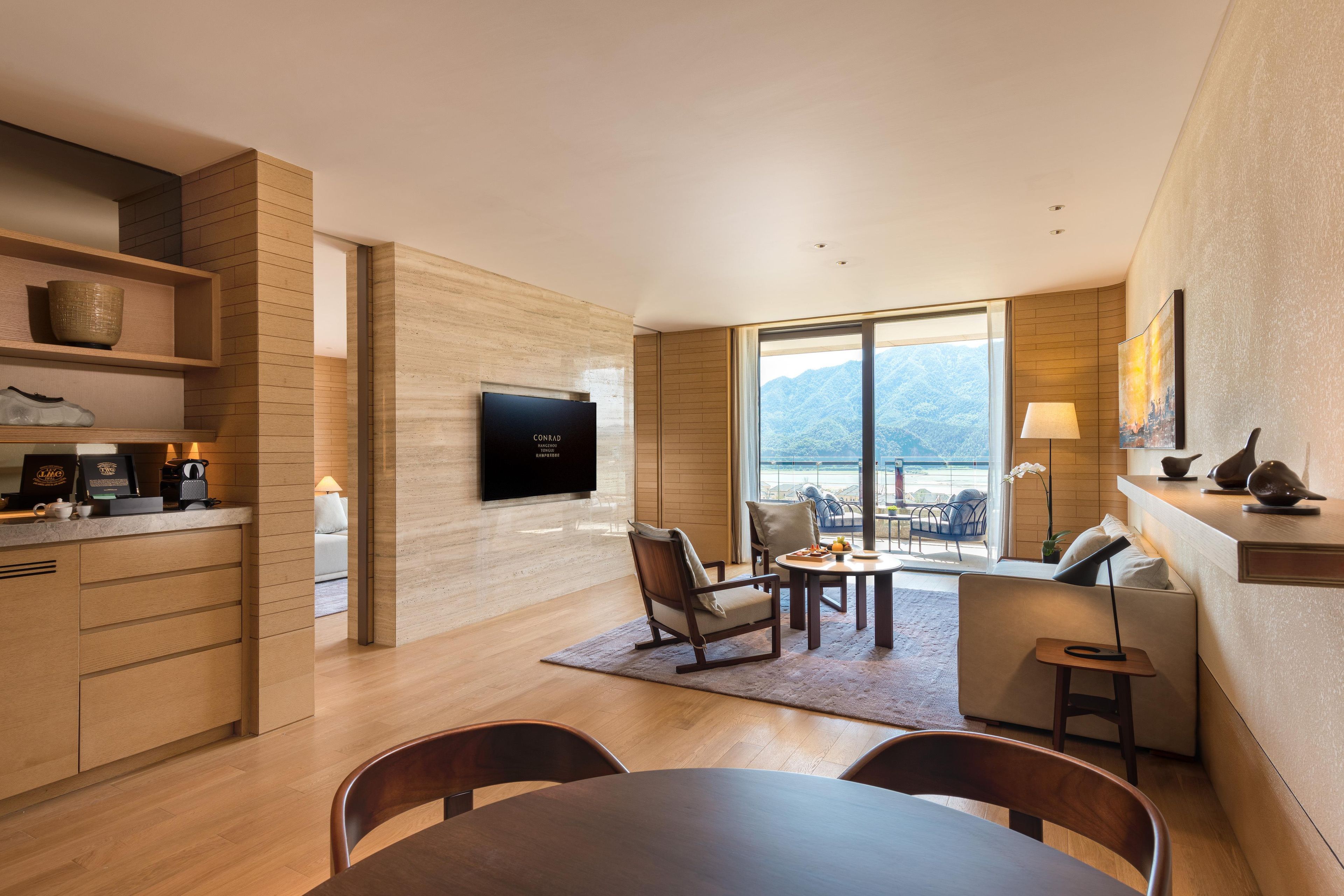 A modern living room with a flat-screen TV, wooden furniture, a dining area, and a large window offering a mountain view.