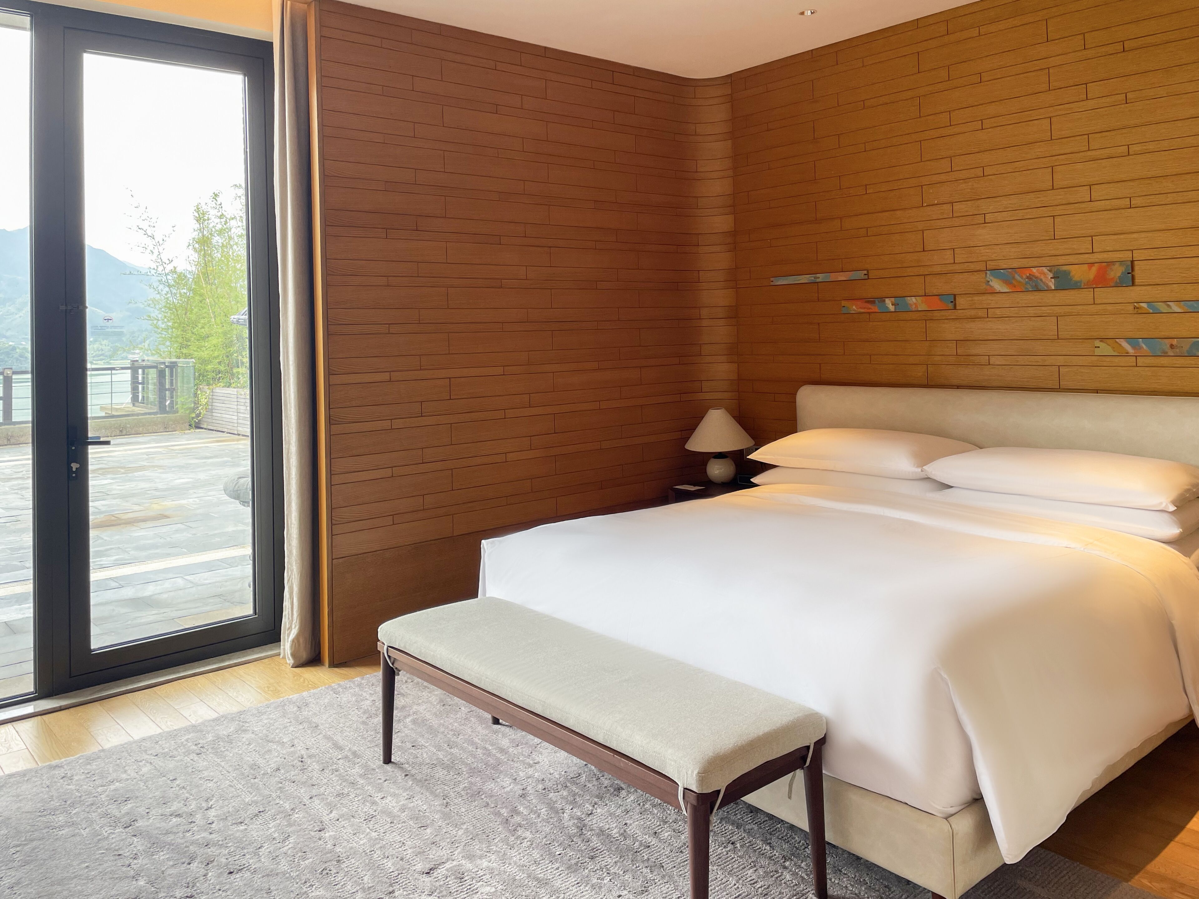 A modern hotel room with a large bed, a bench, a nightstand with a lamp, and a view of the outdoors through a sliding door.