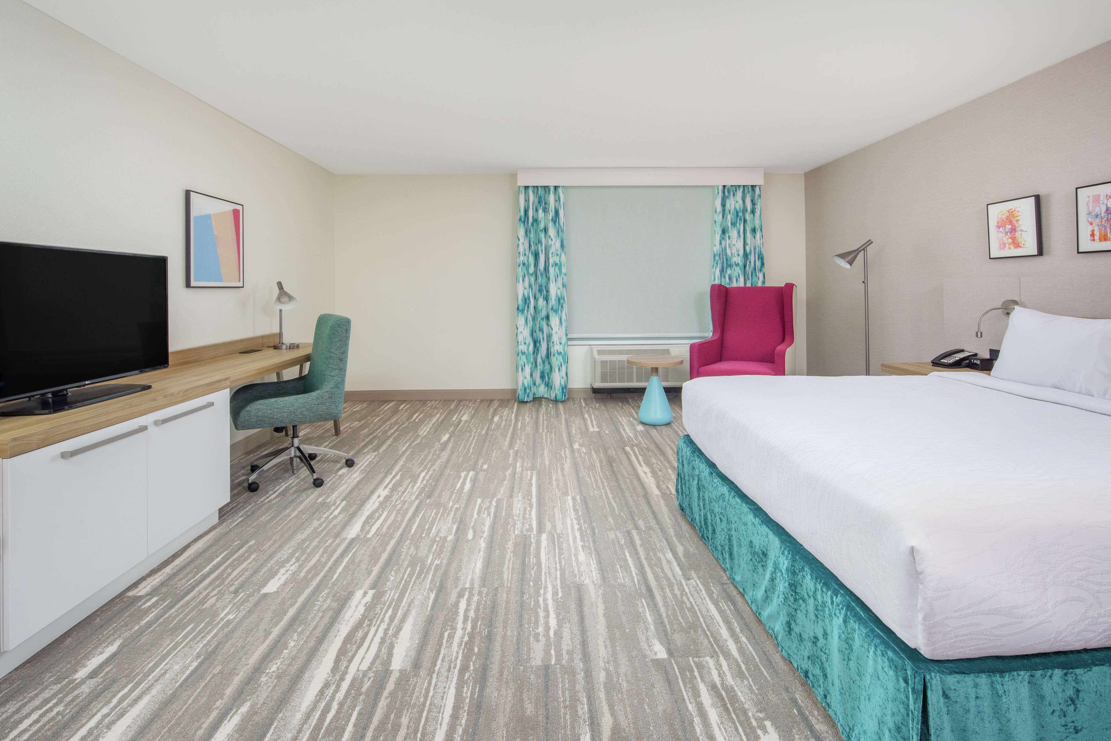 A hotel room with a bed, a desk with a TV, a chair, a desk lamp, a nightstand, and a window with curtains.