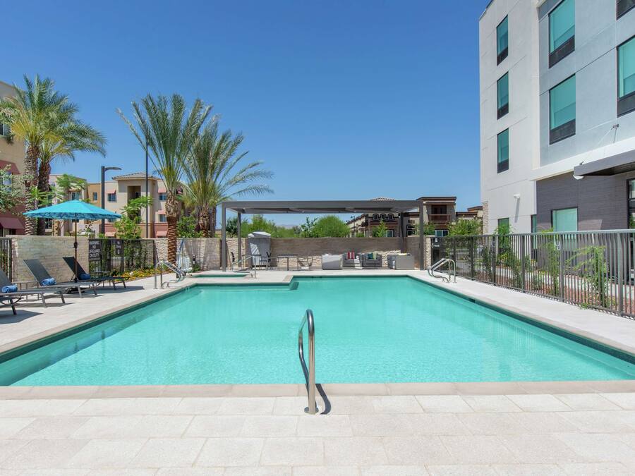 A rectangular swimming pool with a metal handrail, surrounded by lounge chairs and umbrellas, with buildings and palm trees in the background.