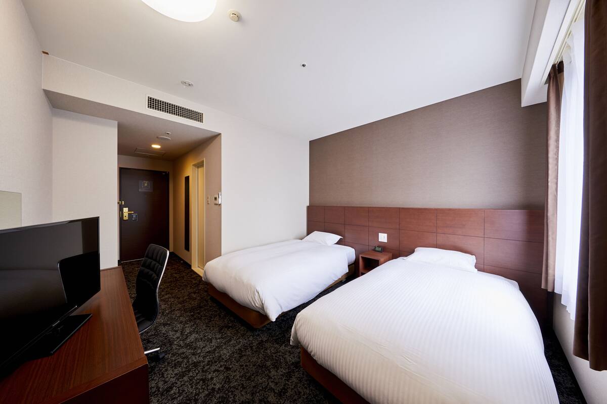 A hotel room with two beds, a desk, a TV, and a door leading to another room.