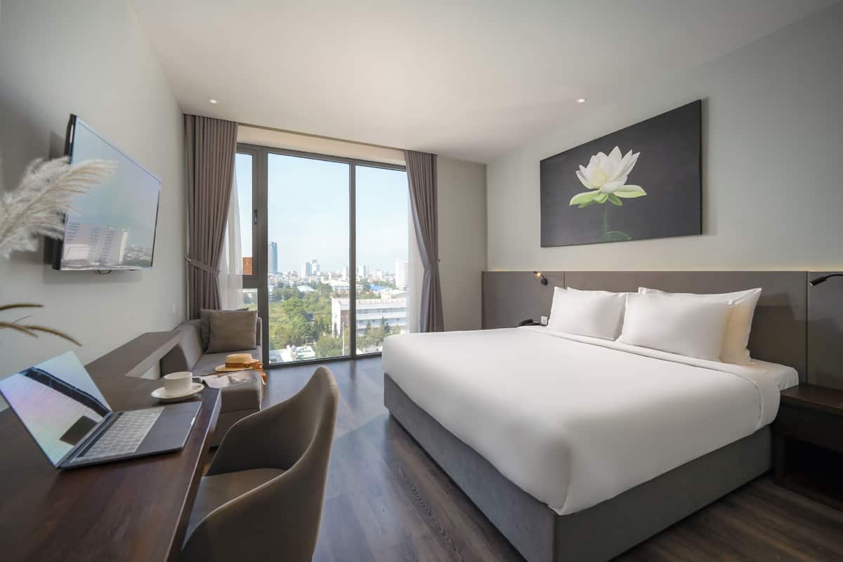 A modern hotel room with a large bed, a desk with a laptop, a chair, and a view of the city.