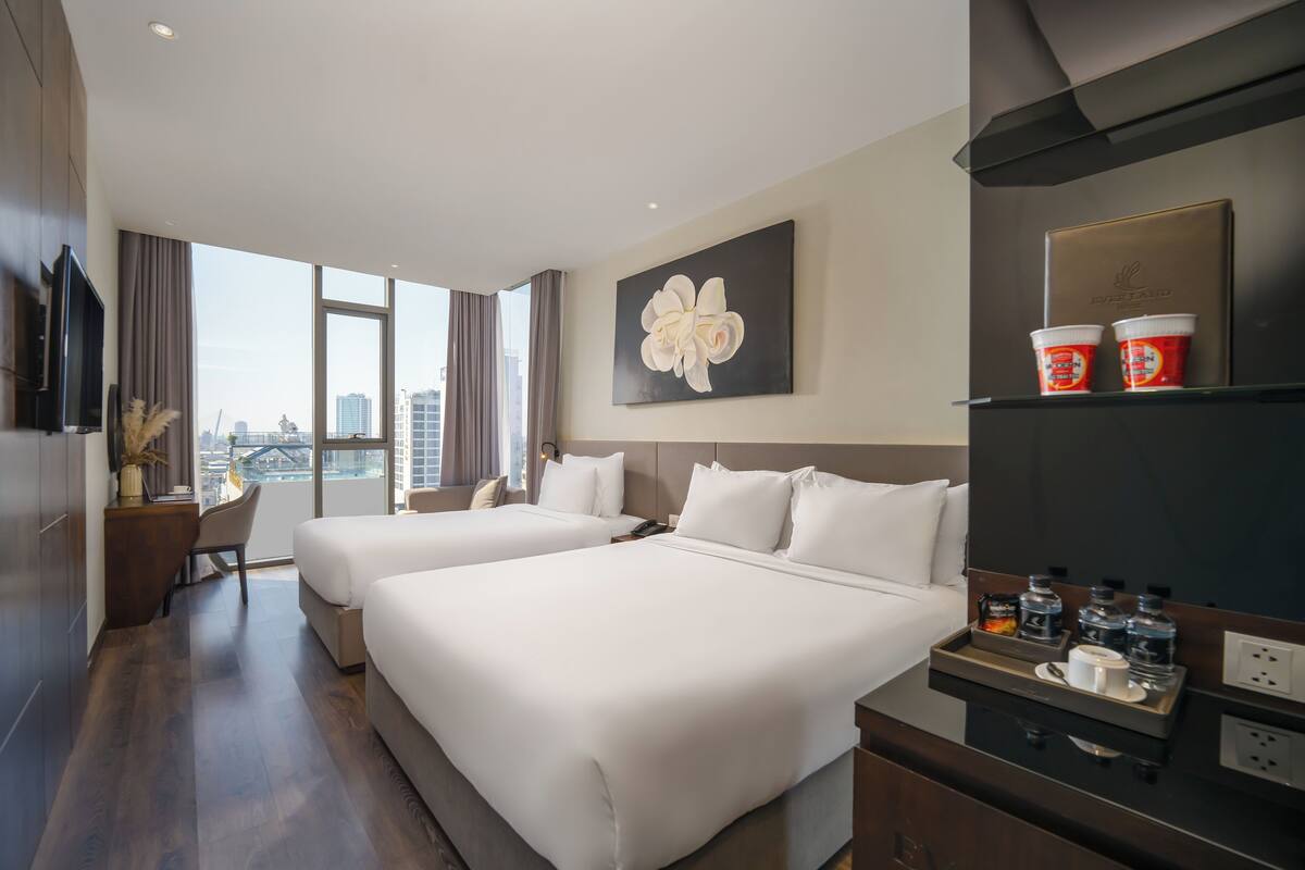 A hotel room with two beds, a desk, a chair, a large window with a city view, and a painting on the wall.