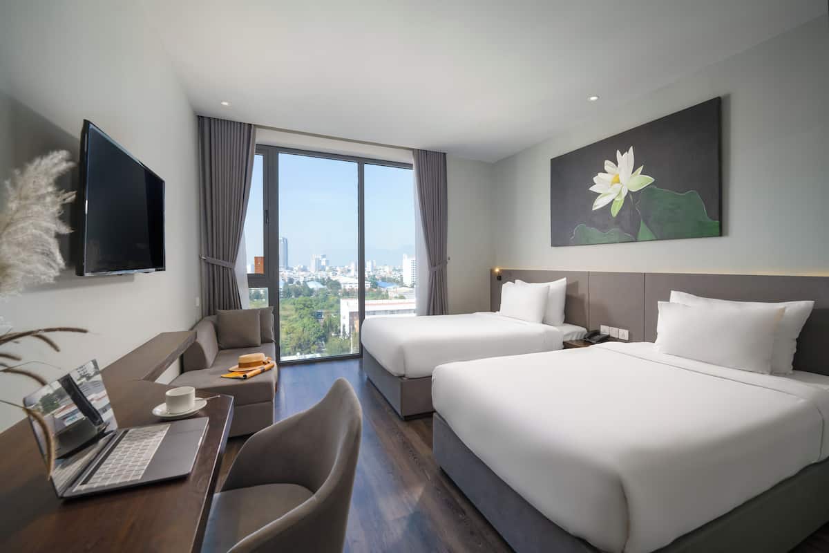 A hotel room with a large window, two beds, a desk with a laptop, a TV, and a painting of a flower.