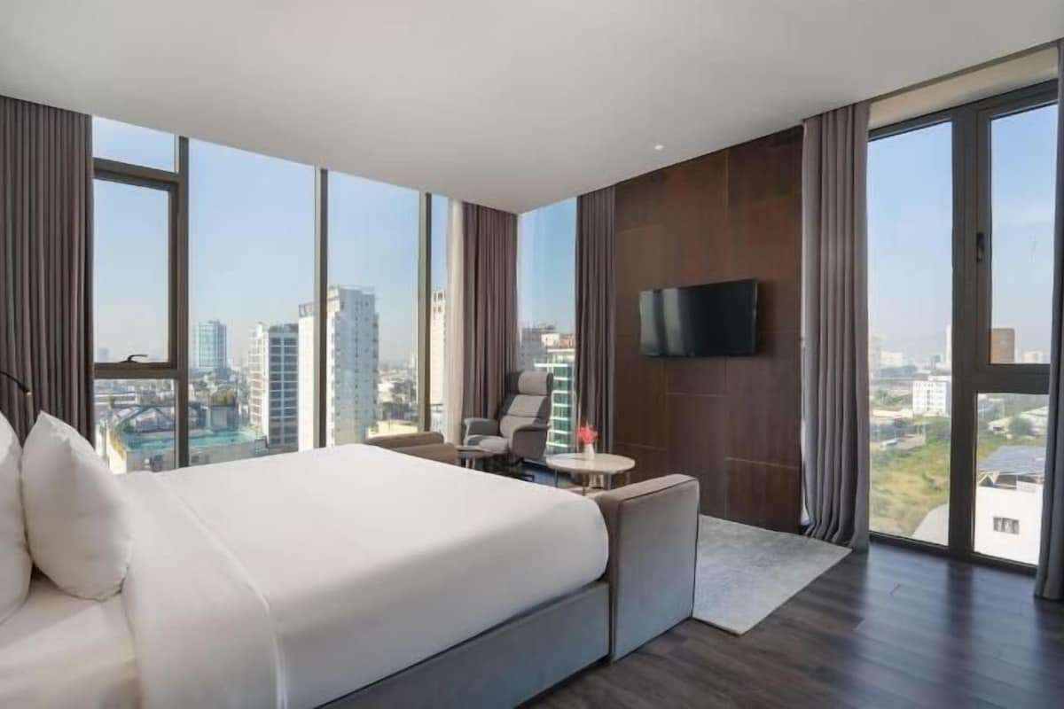 Un moderno cuarto de hotel con una cama grande, un televisor montado en la pared y vistas de la ciudad a través de amplios ventanales.