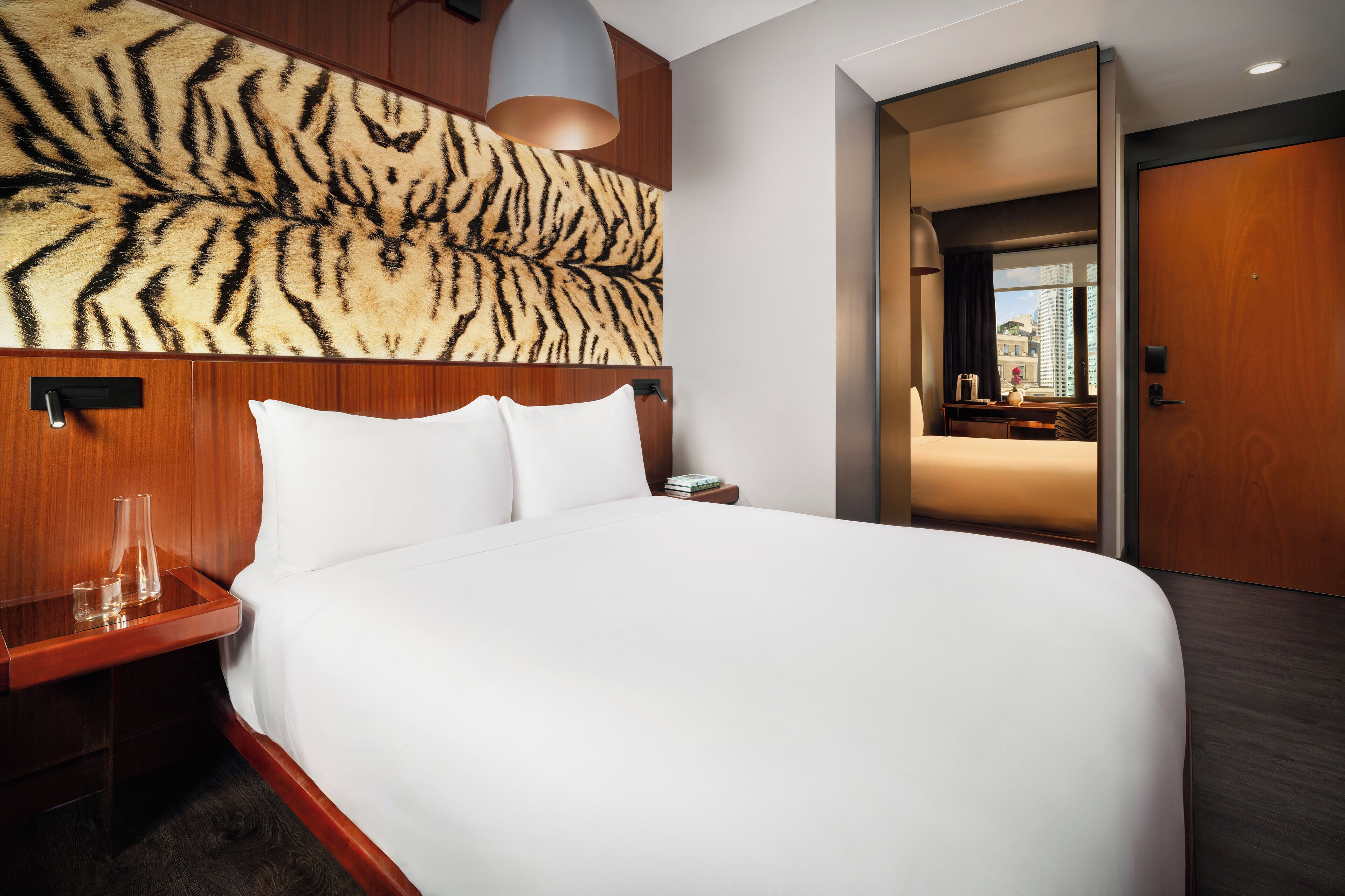 Een hotelkamer met een groot bed, een houten hoofdbord, een nachtkastje met een lamp, en uitzicht op de stad door een raam.