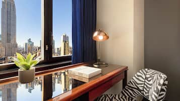 Een kamer met een bureau, een lamp, een fauteuil met zebraprint, een plant en uitzicht op een stadsgezicht met een hoog gebouw.