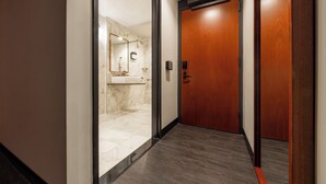 A hallway with a wooden door, a marble-tiled shower area, and a light fixture on the wall.