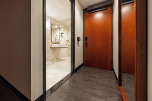 A hallway with a wooden door, a marble-tiled shower area, and a light fixture on the wall.