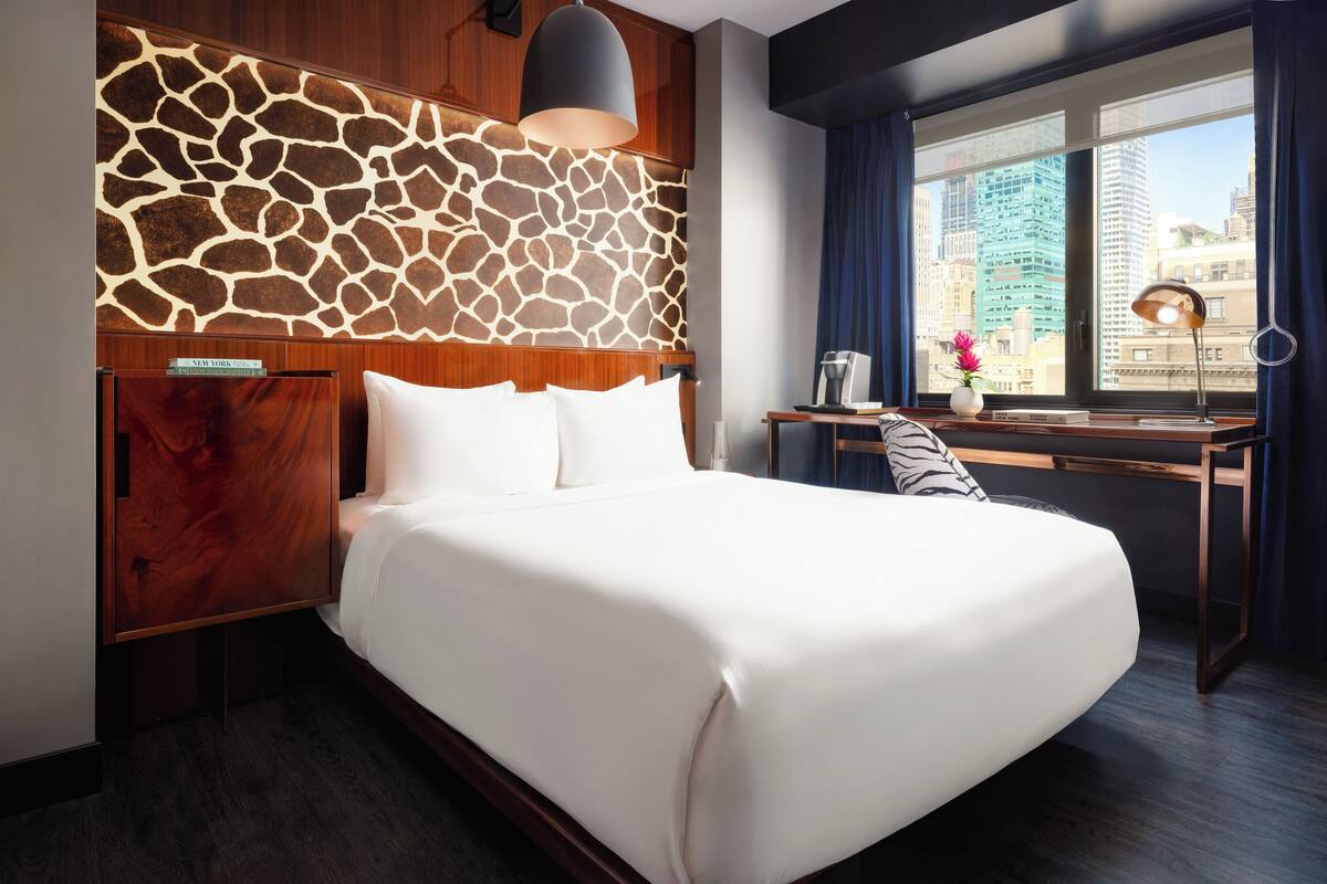 Hotellihuone, jossa on suuri sänky, työpöytä lampun kanssa, ikkunasta avautuva näkymä kaupunkiin ja seinä, jossa on kirahvikuvio.
