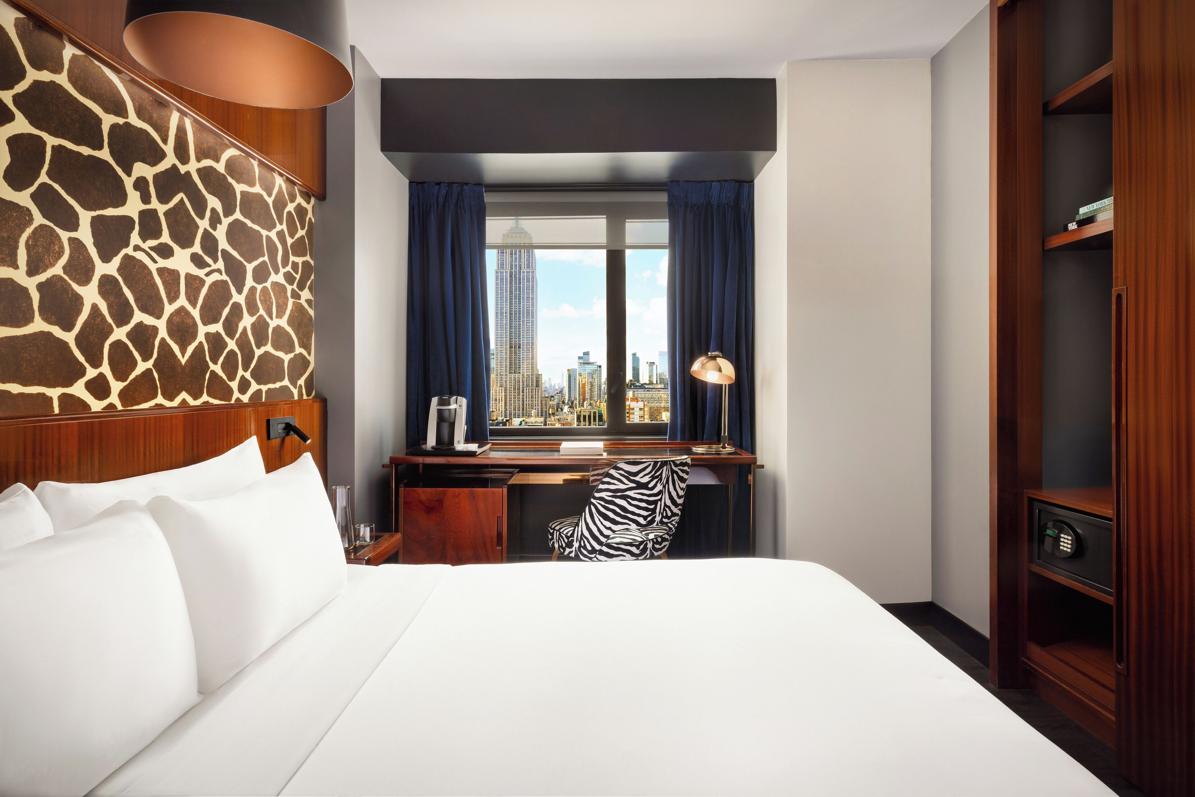Une chambre d’hôtel avec un grand lit, un bureau donnant sur la ville, une chaise à motifs zébrés et une armoire en bois.