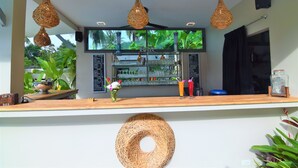 Un área de bar con luces colgantes de mimbre, un ventanal con vistas a la vegetación y un estante con diversos objetos.