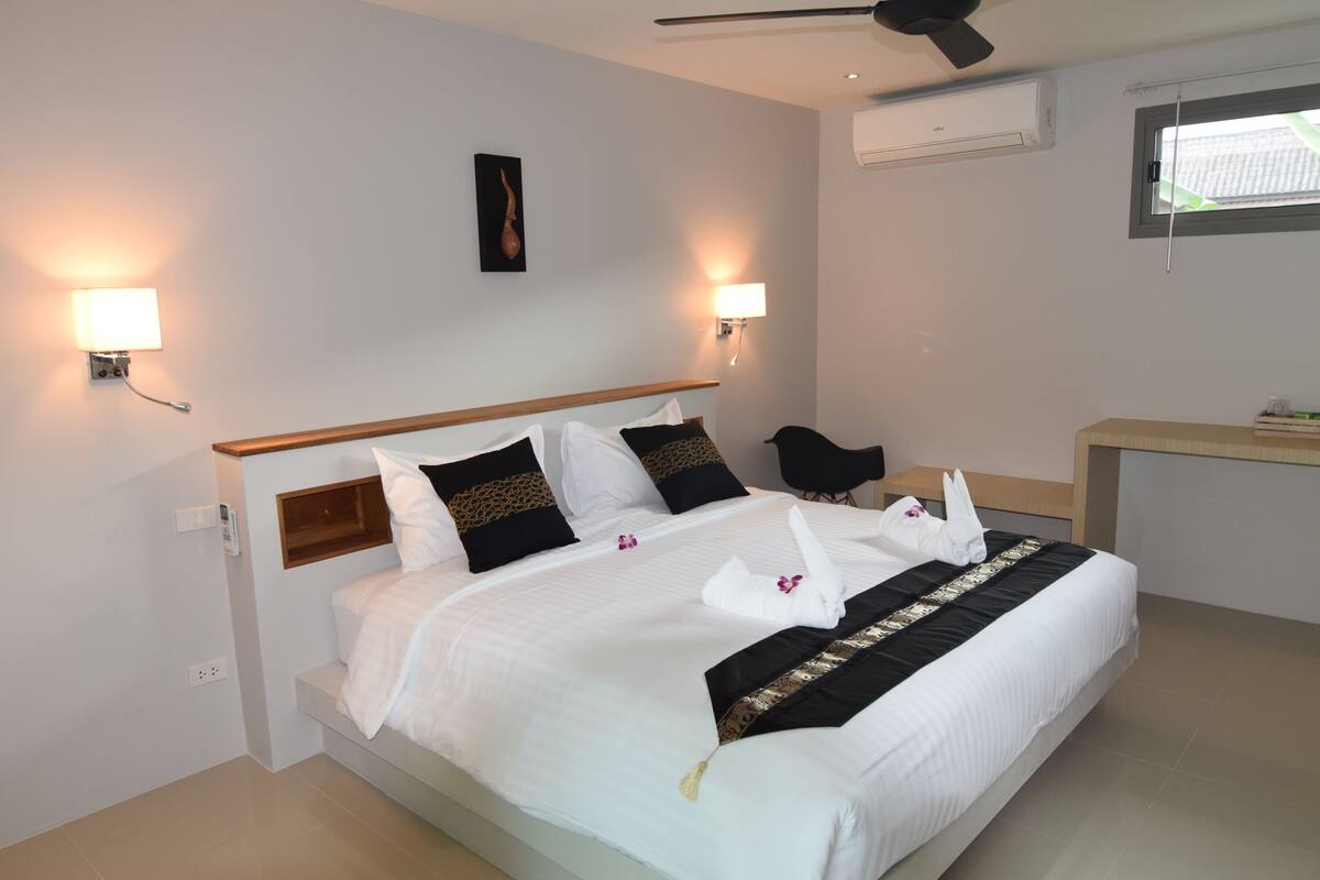 A hotel room with a neatly made bed, two bedside lamps, a small desk, and an air conditioning unit.