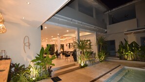 Un área de piscina al aire libre con zona de comedor, rodeada de plantas en macetas y un edificio moderno con balcón.