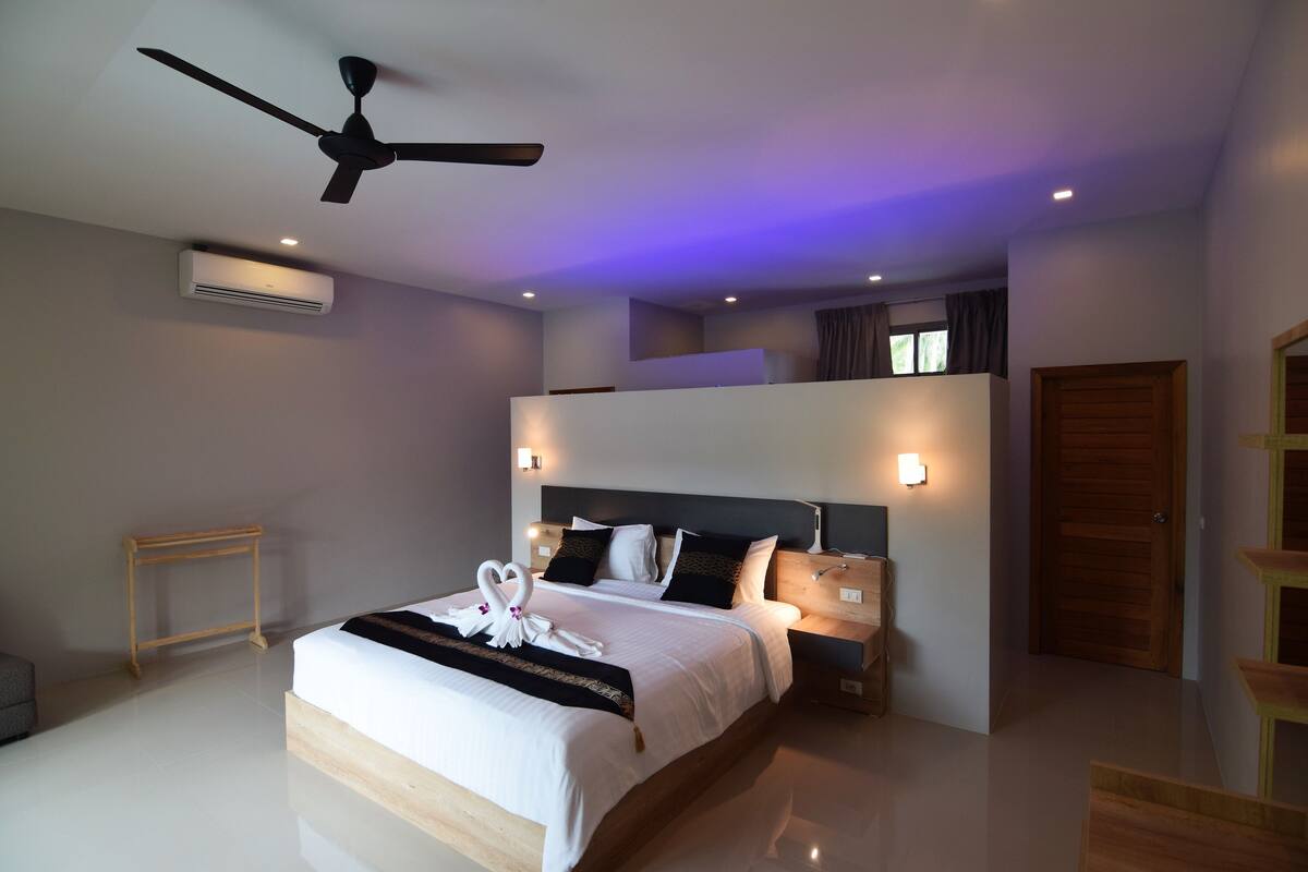 A modern hotel room with a large bed, a ceiling fan, an air conditioning unit, and a wooden nightstand.