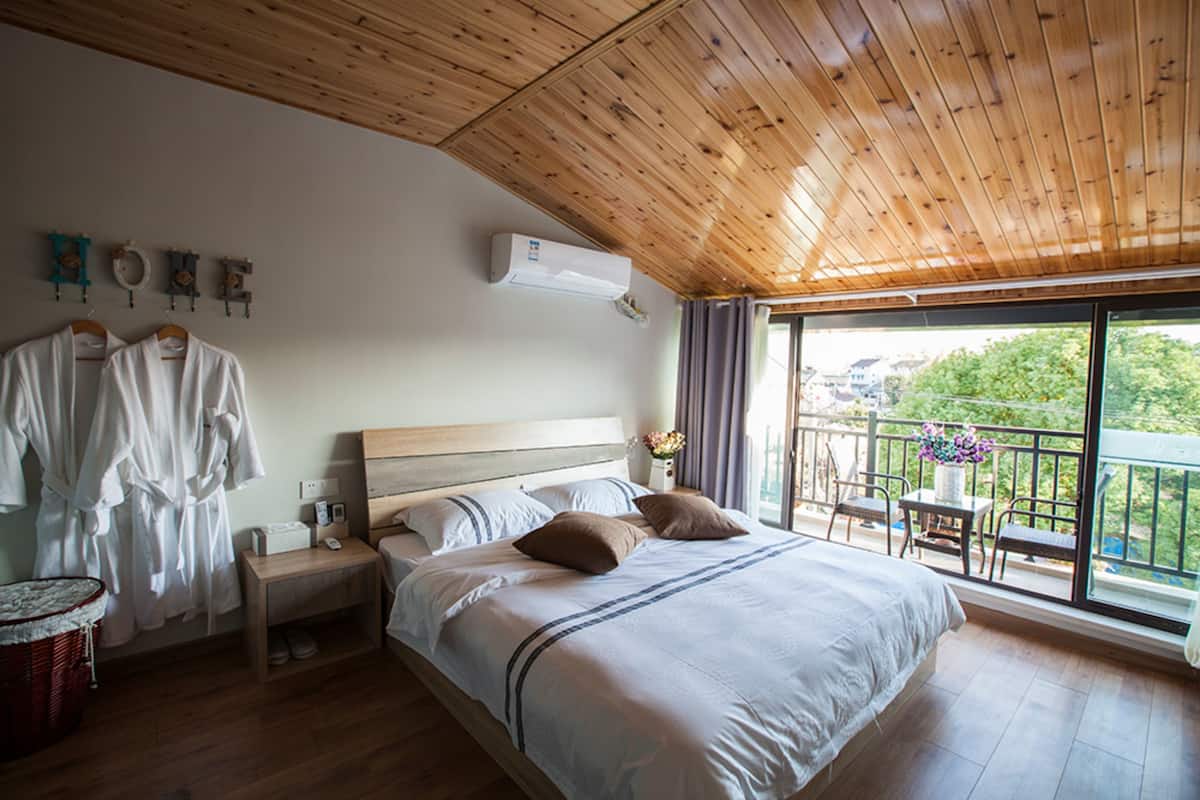 Ein Schlafzimmer mit Holzboden, einem großen Bett mit Kissen, einem Nachttisch, einem Korb und Blick ins Freie vom Balkon aus.